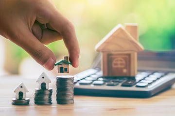 Hand placing a small figurine of a home on top of a stack of coins on a table with a calculator